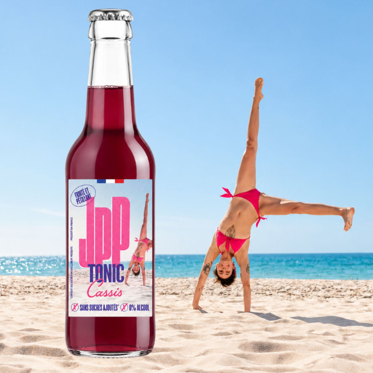 Bouteille de JPP Tonic Cassis sur une plage, une femme fait la roue au bord de l'eau